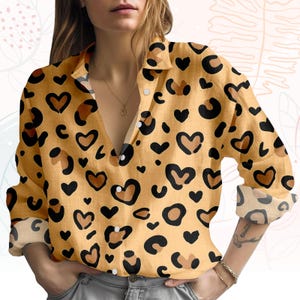May include: A button-down shirt with a yellow background and a pattern of black and brown hearts and leopard spots. The long-sleeved shirt has a collared neckline and rolled-up sleeves.