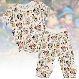 May include: Cream-coloured children's pyjama set with a repeating pattern of Mickey Mouse, Minnie Mouse, Donald Duck, Daisy Duck, Goofy, and Pluto in safari outfits. The set includes a short-sleeved top and matching trousers.