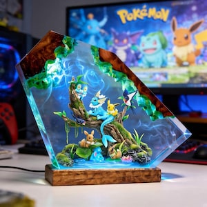 May include: A custom resin and wood lamp featuring a diorama of multiple small Pokemon figures, including Vaporeon, Eevee, and Froakie, arranged on a textured tree branch and mossy base. The piece emits a bright blue light through the clear resin.