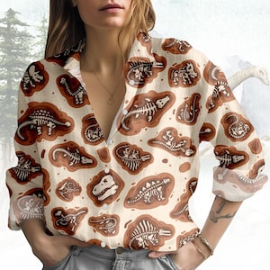 May include: A button-down shirt with a beige background and a repeating pattern of brown dinosaur fossils. The long-sleeved shirt has a collared neckline and is made of a lightweight fabric.
