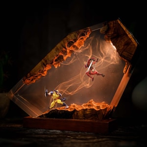 Op de afbeelding: Een decoratieve lamp bestaande uit een helder harsblok met natuurlijke houten randen. Binnenin de hars zijn miniatuurfiguren van Wolverine in een geel-blauw pak en Deadpool in een rood pak opgehangen, verlicht door een oranje gloed.