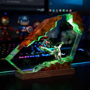 May include: A rectangular resin and wood display piece featuring a miniature scene of a superhero in a red and blue suit battling a green reptilian creature. The clear resin contains glowing green swirls and is mounted on a wooden base.