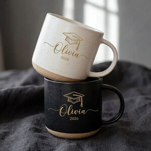 May include: Two ceramic mugs, one white and one black, stacked. The white mug features a graduation cap graphic and the name "Olivia 2026" in gold script. The black mug has the same design. Both mugs have a speckled texture and a beige base.