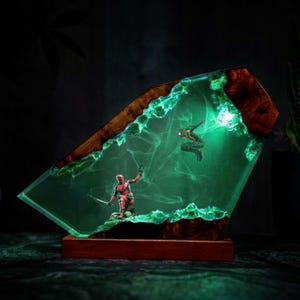 Könnte beinhalten: Eine dekorative Lampe mit einem Holzrahmen und einem Kern aus durchscheinendem grünem Harz. Im Harz kniet eine rote Deadpool-Figur mit einem Schwert, während eine schwarze Spider-Man-Figur in einer dynamischen Pose schwebt. Das Objekt leuchtet grün.