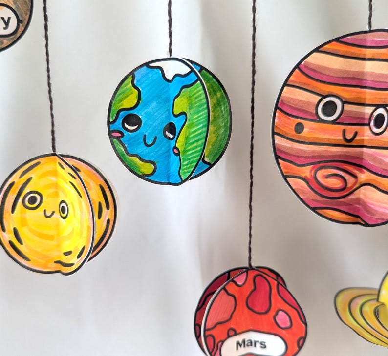 May include: A hanging mobile featuring several paper planets with hand-drawn, colorful designs and cute faces. The planets include a blue and green Earth, a yellow planet, a striped orange planet, and a red planet labeled with the text Mars.