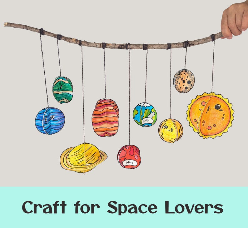 May include: A solar system mobile featuring hand-drawn, colorful paper planets and a sun suspended from a natural wooden branch by black strings. The planets include Earth and Mars, which are labeled with their names in black text.
