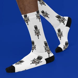 Puede incluir: Calcetines blancos con un patrón repetido de micrófonos de estudio grises y negros. Los calcetines tienen talones y punteras negras. El fondo es azul liso.