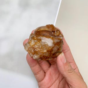 Pode incluir: Uma escultura de cristal esculpida, de cor âmbar. O cristal tem um exterior áspero e texturizado com um topo liso e arredondado. A escultura é segurada em uma mão, mostrando seus detalhes intrincados e variações naturais de cor e clareza.
