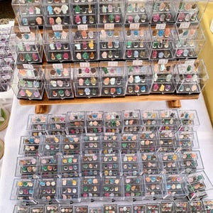 Może przedstawiać: Ekspozycja różnych kolorowych kamieni szlachetnych i minerałów, każdy umieszczony w indywidualnych, przezroczystych plastikowych gablotach. Gabloty są ustawione na drewnianej półce i białej powierzchni, prezentując różnorodne kształty i kolory.