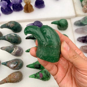 Könnte beinhalten: Eine Hand hält einen geschnitzten grünen Edelstein-Adlerkopf. Der Adlerkopf ist detailliert mit einem gebogenen Schnabel und strukturierten Federn. Im Hintergrund sind weitere geschnitzte Vogelköpfe aus Stein in verschiedenen Farben zu sehen.