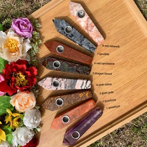 May include: An assortment of eight gemstone pipes in various colors and patterns, including red network, larvikite, red sandstone, dragon blood, pink zebra, gold jade, sunstone, and amethyst. Each pipe has a metal bowl and is displayed on a wooden tray.