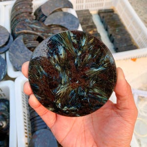 May include: A round, polished stone with a dark brown base and intricate blue and gold crystalline patterns. The stone is held in a hand, showcasing its unique texture and color variations. Other similar stones are visible in the background.