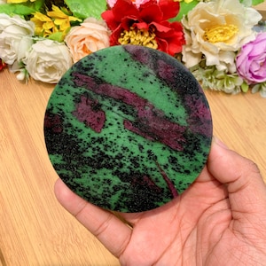 May include: A circular stone coaster with a green base, featuring dark red and black veining. The coaster is held in a hand, with a blurred floral arrangement in the background.