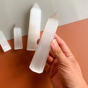 May include: A collection of white selenite crystal wands of different sizes, set against a two-tone backdrop. One wand is held in a hand. The wands are smooth, polished, and translucent, each with a pointed top.