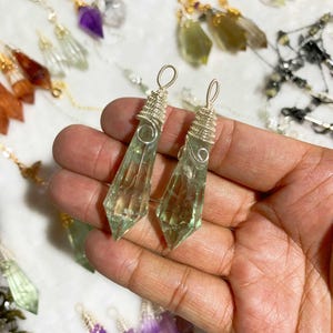 May include: Two light green crystal pendants with silver wire wrapping are held in a hand. The pendants are elongated and faceted, with a pointed bottom. Other crystal pendants in various colors, including purple and clear, are visible in the background.