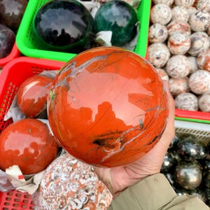 Könnte beinhalten: Eine polierte, leuchtend orange-rote Kugel mit natürlichen Adern und Mustern. Die Edelsteinkugel wird in einer Hand gehalten, wobei im Hintergrund weitere Kugeln in verschiedenen Farben und Texturen zu sehen sind, die in roten und grünen Körben ausgestellt sind.