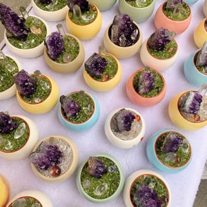 Könnte beinhalten: Eine Sammlung kleiner, farbenfroher Keramiktöpfe, die jeweils einen Cluster aus violetten Amethystkristallen und klaren QuarzKristallen enthalten. Die Töpfe sind mit grünem Moos und kleinen dekorativen Steinen gefüllt. Die Töpfe sind in verschiedenen Farben erhältlich, darunter Gelb, Rosa und Blau.