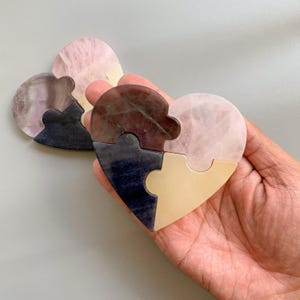 May include: A heart-shaped jigsaw puzzle composed of four pieces in pink, purple, navy, and beige. The puzzle is held in a hand, with the pieces interlocked to form a complete heart shape. The background is a plain grey.