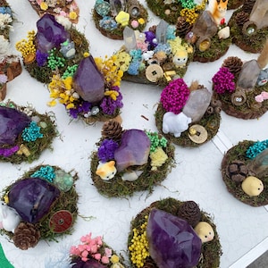 Könnte beinhalten: Eine Sammlung dekorativer Kristall- und Moosarrangements. Jedes Arrangement zeigt einen großen, polierten Amethystkristall, umgeben von Moos, Trockenblumen, Miniaturfiguren und Tannenzapfen. Die Arrangements sind auf einer weißen Oberfläche ausgestellt.