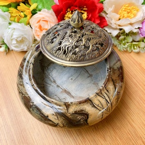 Peut inclure: Un brûleur d'encens décoratif avec couvercle. Le brûleur est en pierre marbrée aux tons marron, beige et gris. Le couvercle est de couleur bronze et présente un motif de dragon. Le brûleur est rond et a une finition lisse et polie.