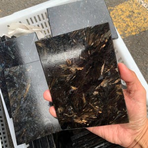 Op de afbeelding: Een rechthoekige, gepolijste zwarte steen met een opvallend patroon van goudbruine, naaldachtige insluitsels. De steen wordt in een hand gehouden, met andere soortgelijke stenen zichtbaar op de achtergrond, wat wijst op een verzameling.