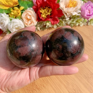 May include: Two black and red gemstone spheres, also known as red aventurine, are shown in a hand. The spheres are smooth and polished.