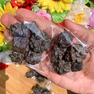 May include: A close-up of a bag of dark grey, crystal-like stones. The stones are in a clear plastic bag and are being held by a hand.