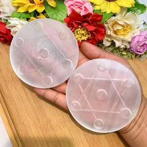May include: Two round, translucent selenite crystal plates. Each plate features a six-pointed star design with raised circular accents. The plates are displayed against a backdrop of colorful flowers and a wooden surface.