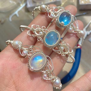 Könnte beinhalten: Handgefertigte Armbänder aus Silberdraht mit ovalen und runden Edelsteinen und kleinen Perlen. Zu den Edelsteinen gehören ein blauer, ein klarer und ein regenbogenfarbener Stein. Die Armbänder werden in einer Hand gehalten.