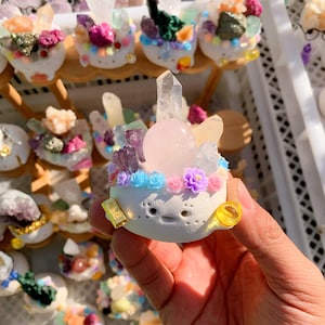 Puede incluir: Una figura de cerámica blanca adornada con una variedad de cristales, incluyendo una esfera de cuarzo rosa y puntas de cuarzo transparente. La figura está decorada con pequeñas flores coloridas. Otras figuras similares son visibles en el fondo.