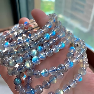 May include: Several strands of beaded bracelets. The beads are round, translucent, and gray, with flashes of blue and iridescent color. The bracelets are held in a hand, showcasing the jewelry's design and color variations.