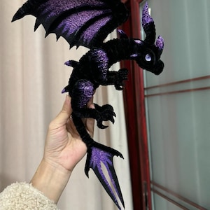 Może przedstawiać: Pluszowy czarny smok z fioletowymi brokatowymi akcentami na skrzydłach, rogach i ogonie. Smok ma szczegółowe, teksturowane ciało i jest ustawiony ze skrzydłami rozłożonymi. To przedmiot o tematyce fantasy.