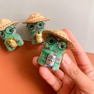 May include: Three green, textured figurines wearing straw hats and round glasses. Each figurine holds a miniature item: a Starbucks cup, a small bottle, and a can with a cartoon character. The figurines have pink accents.