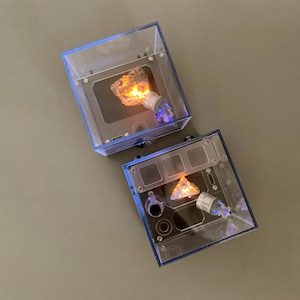 以下が含まれることがあります： 青い縁取りのある、透明な正方形のディスプレイケースが2つ。各ケースには、琥珀色の光る岩と小さな透明なライトが入っています。ケースは灰色の表面にあります。