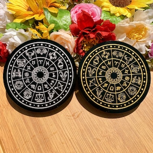 May include: Two black round coasters with gold zodiac symbols and white outlines. The coasters feature the twelve signs of the zodiac in a circular pattern.