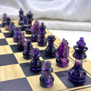 May include: A chess set with translucent purple and black pieces on a wooden chessboard. The pieces include pawns, rooks, knights, bishops, a queen, and a king. The board has black and light brown squares.