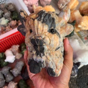 May include: A carved stone figurine of a bull with black and tan coloring. The bull has small horns and a detailed face. The figurine is held in a hand, with other similar figurines visible in the background. The bull is a decorative item.