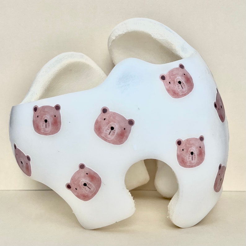 May include: White helmet with a pattern of brown bear faces. The helmet is made of soft foam and has a rounded shape.