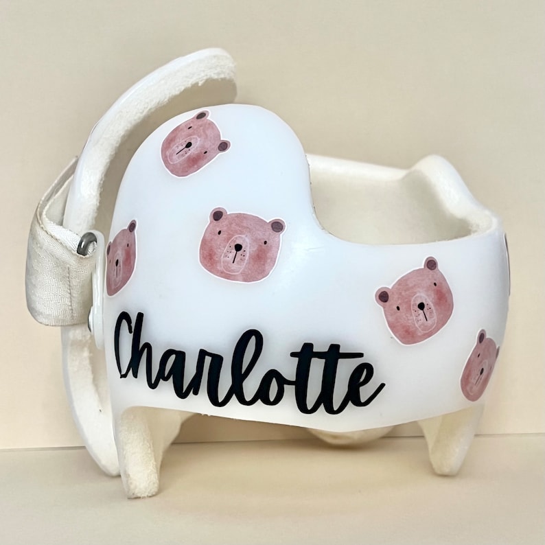 May include: White helmet with a personalized name "Charlotte" in black cursive lettering. The helmet is decorated with multiple brown bear stickers with pink accents.