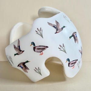 May include: White helmet with a pattern of flying ducks and cattails. The ducks are brown, green, and gray. The cattails are green and brown.