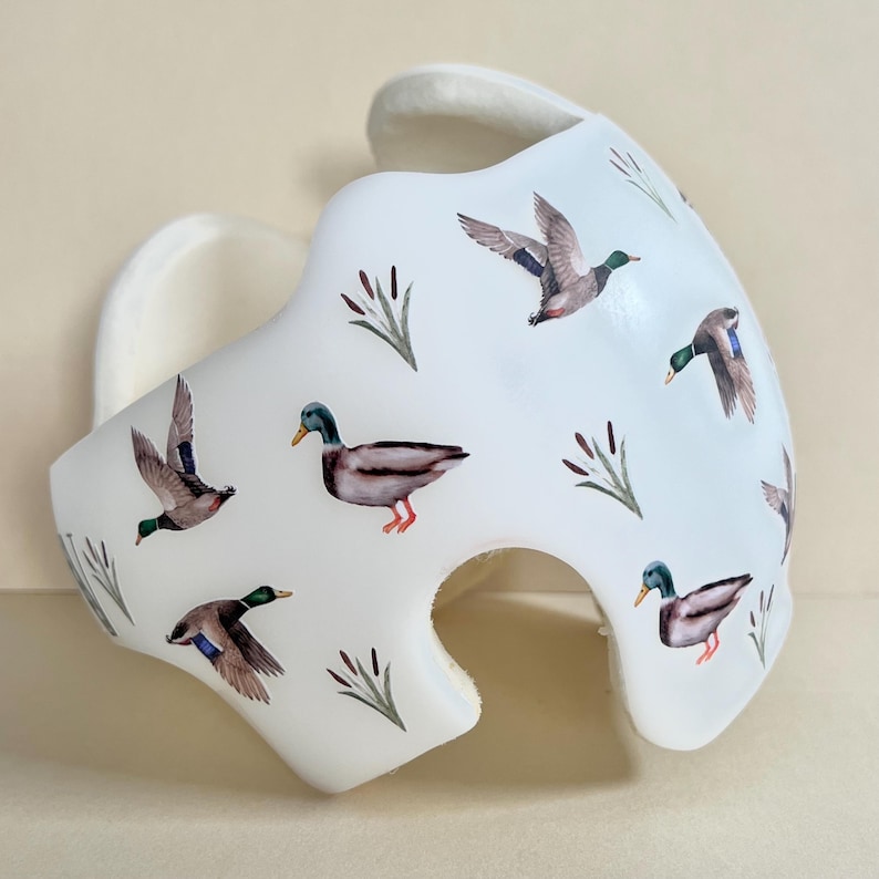 May include: White helmet with a pattern of flying ducks and cattails. The ducks are brown, green, and gray. The cattails are green and brown.