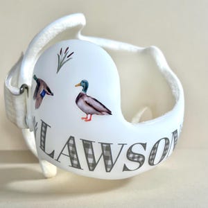 May include: White helmet with a black and white checkered pattern that says "LAWSON" on the side. The helmet has a duck and a floral design on the front.