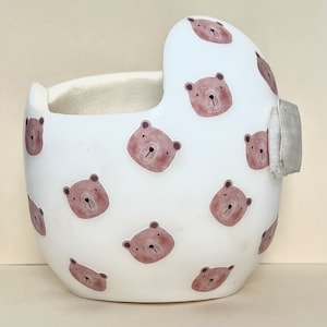 May include: White helmet with a brown bear pattern. The helmet has a soft, padded interior and a smooth, hard exterior. The bear pattern is repeated throughout the helmet.