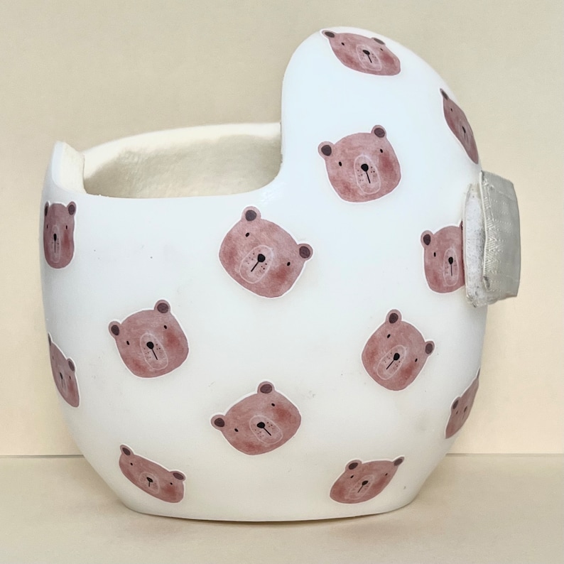 May include: White helmet with a brown bear pattern. The helmet has a soft, padded interior and a smooth, hard exterior. The bear pattern is repeated throughout the helmet.