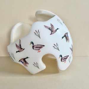 May include: A white helmet with a pattern of ducks and reeds. The helmet is made of foam and has a soft lining.