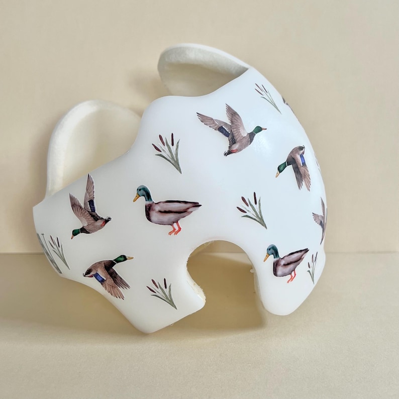 May include: A white helmet with a pattern of ducks and reeds. The helmet is made of foam and has a soft lining.