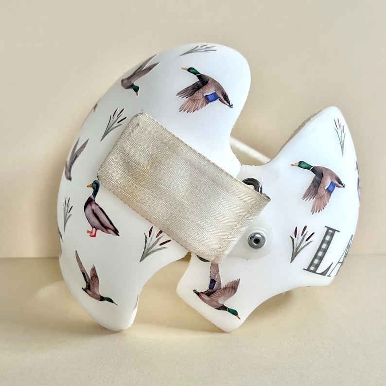 May include: White helmet with a duck pattern and the letter "L" on the side. The helmet has a strap with a white buckle.