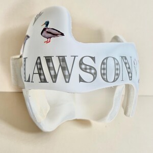 May include: White helmet with a black and white checkered pattern and the name "LAWSON" printed on it. The helmet has a duck design on the side.