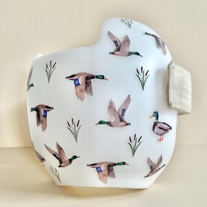 May include: White orthopedic helmet with a pattern of brown and green ducks flying over a white background. The helmet has a beige strap.