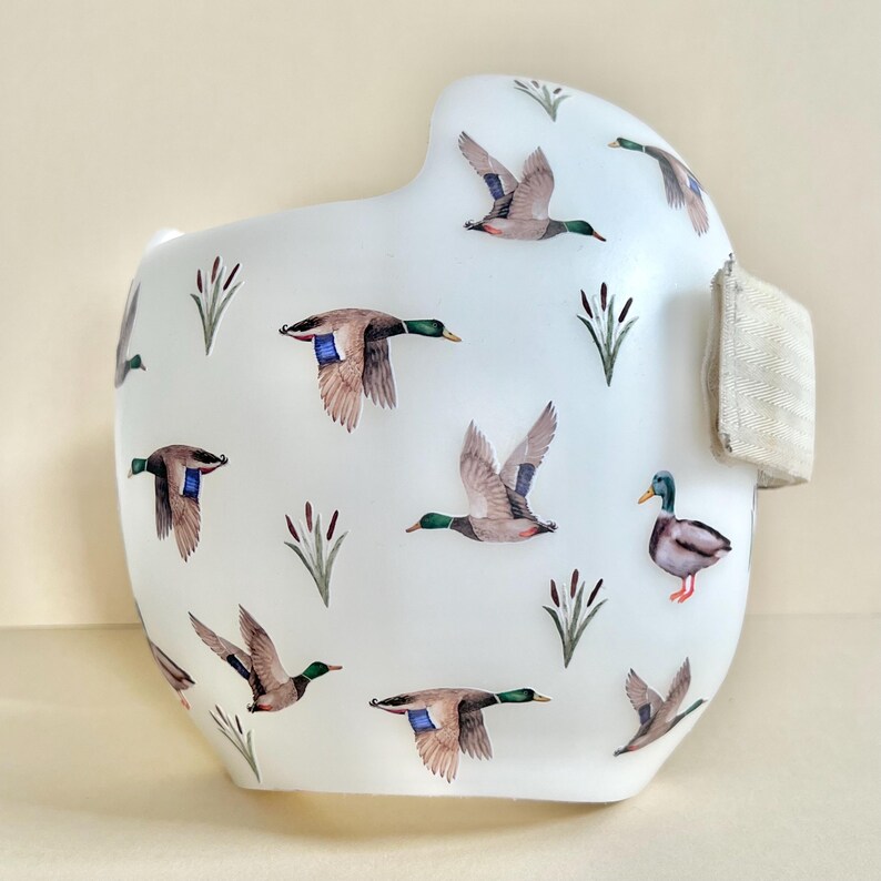 May include: White orthopedic helmet with a pattern of brown and green ducks flying over a white background. The helmet has a beige strap.
